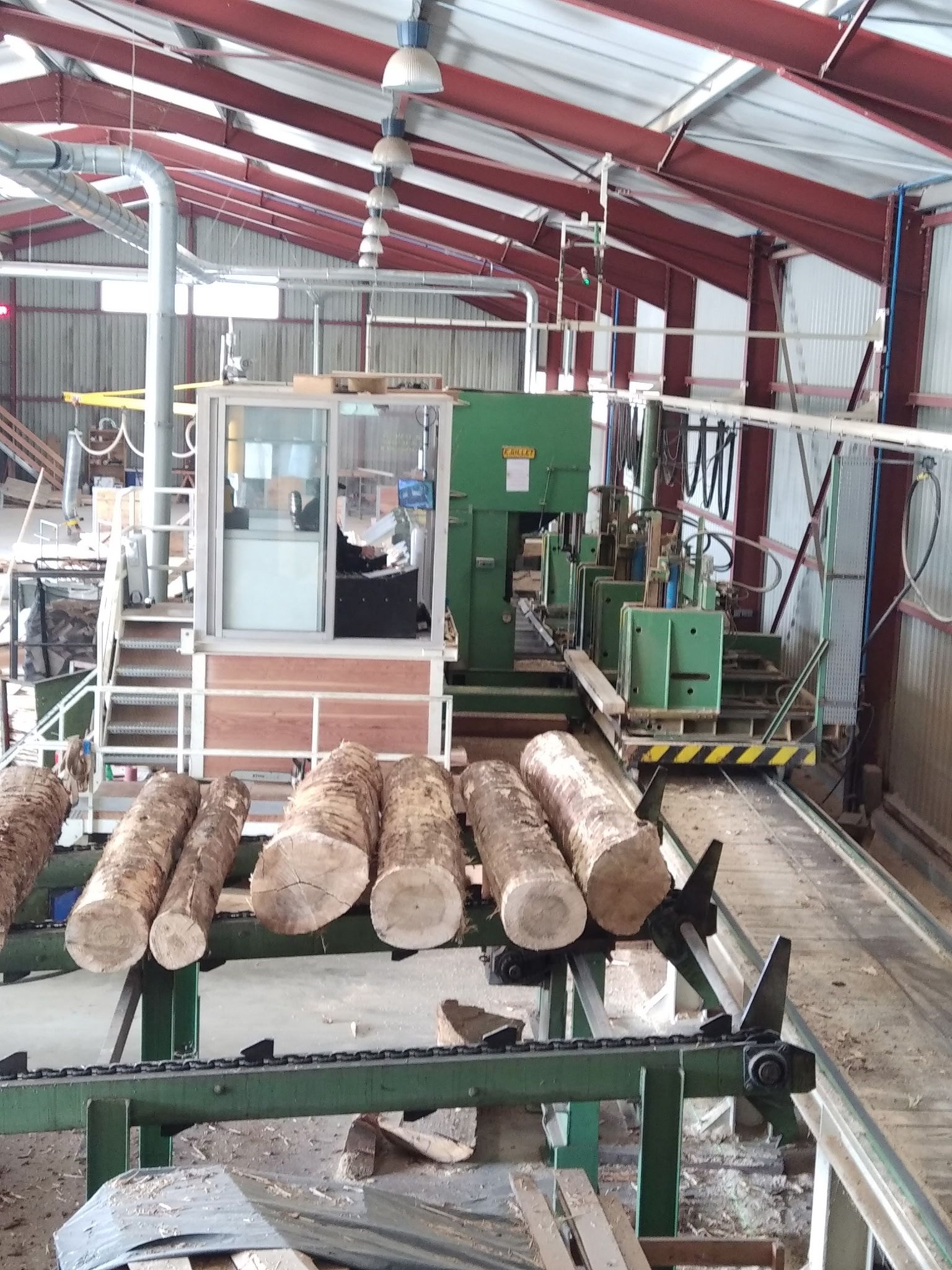 Intérieur d’une scierie équipée de machines industrielles, avec plusieurs troncs de bois alignés sur un convoyeur en métal menant vers une unité de découpe automatisée.