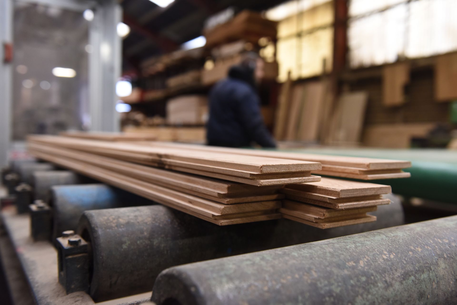 Planches de bois empilées sur des rouleaux de convoyeur dans une scierie, une silhouette floue en arrière-plan.