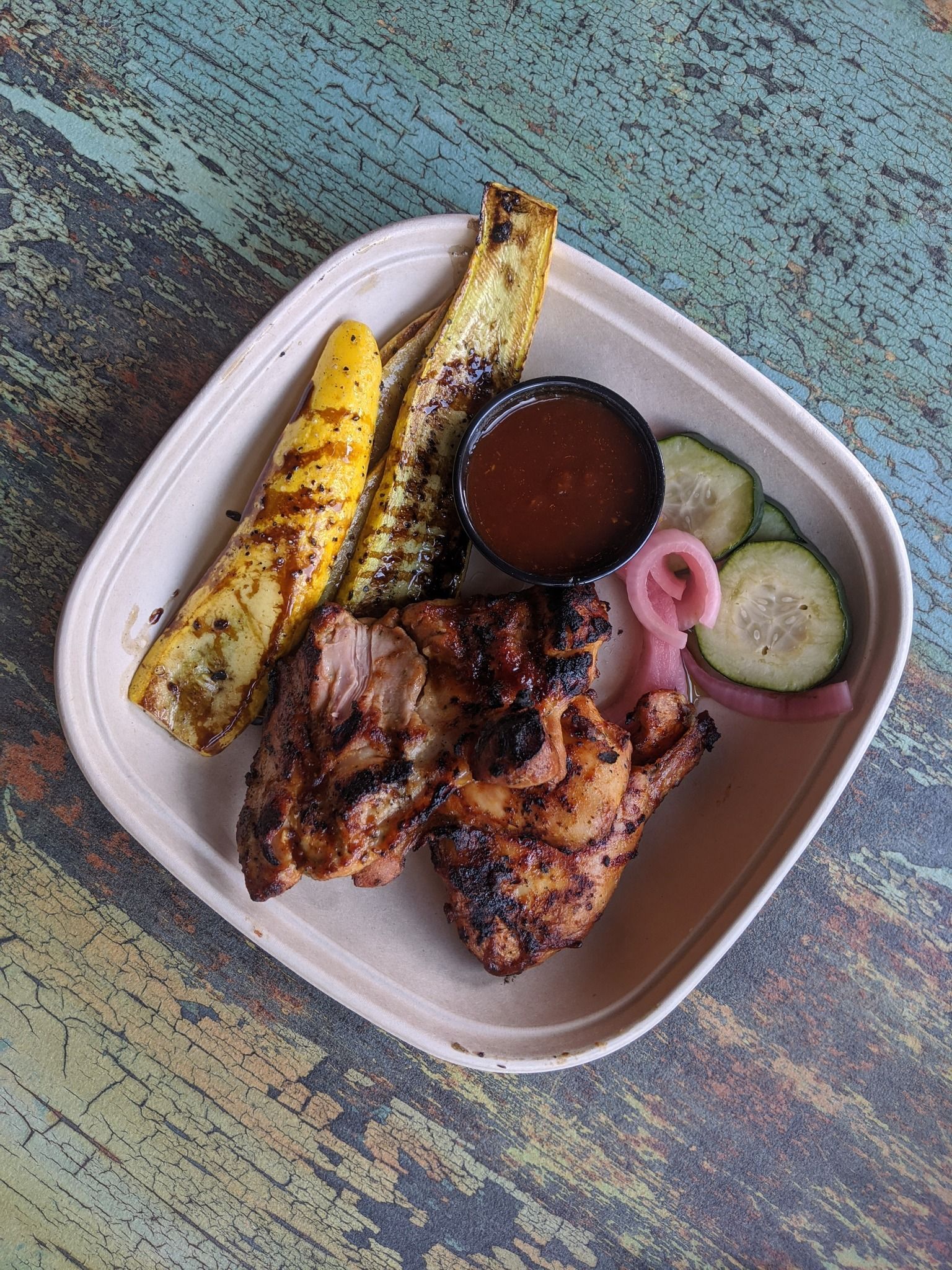 Grilled chicken, squash, sauce, and pickled vegetables in a square container on a patterned surface.