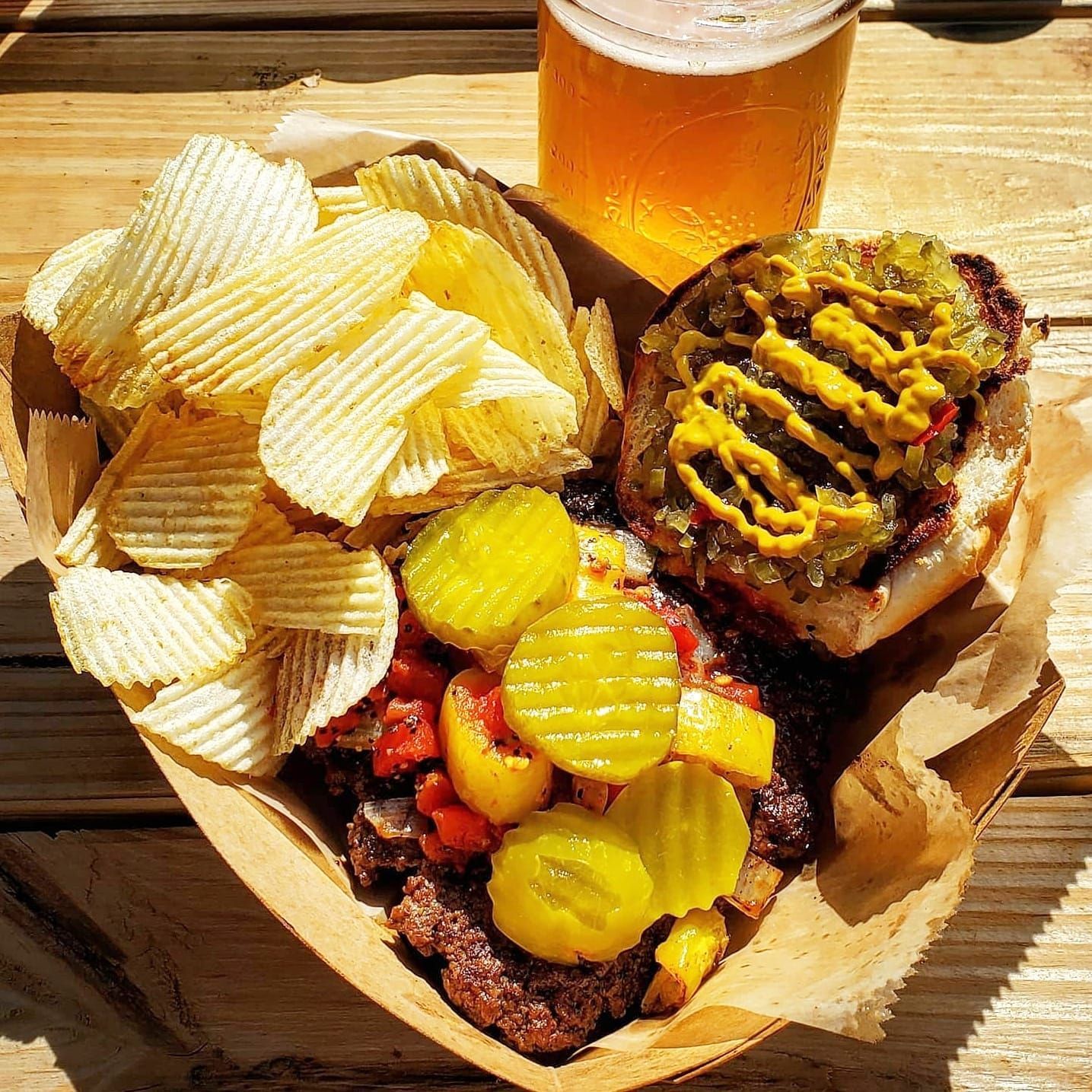 Burger with chips and pickles, with beer in a rustic outdoor setting.