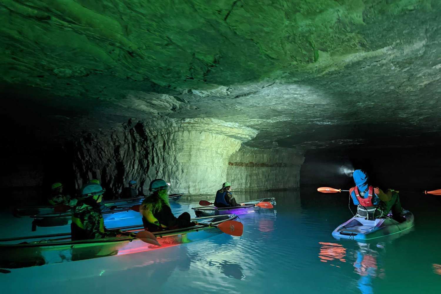 Kayakers with headlamps in an underground cavern with aqua water and colored lighting.