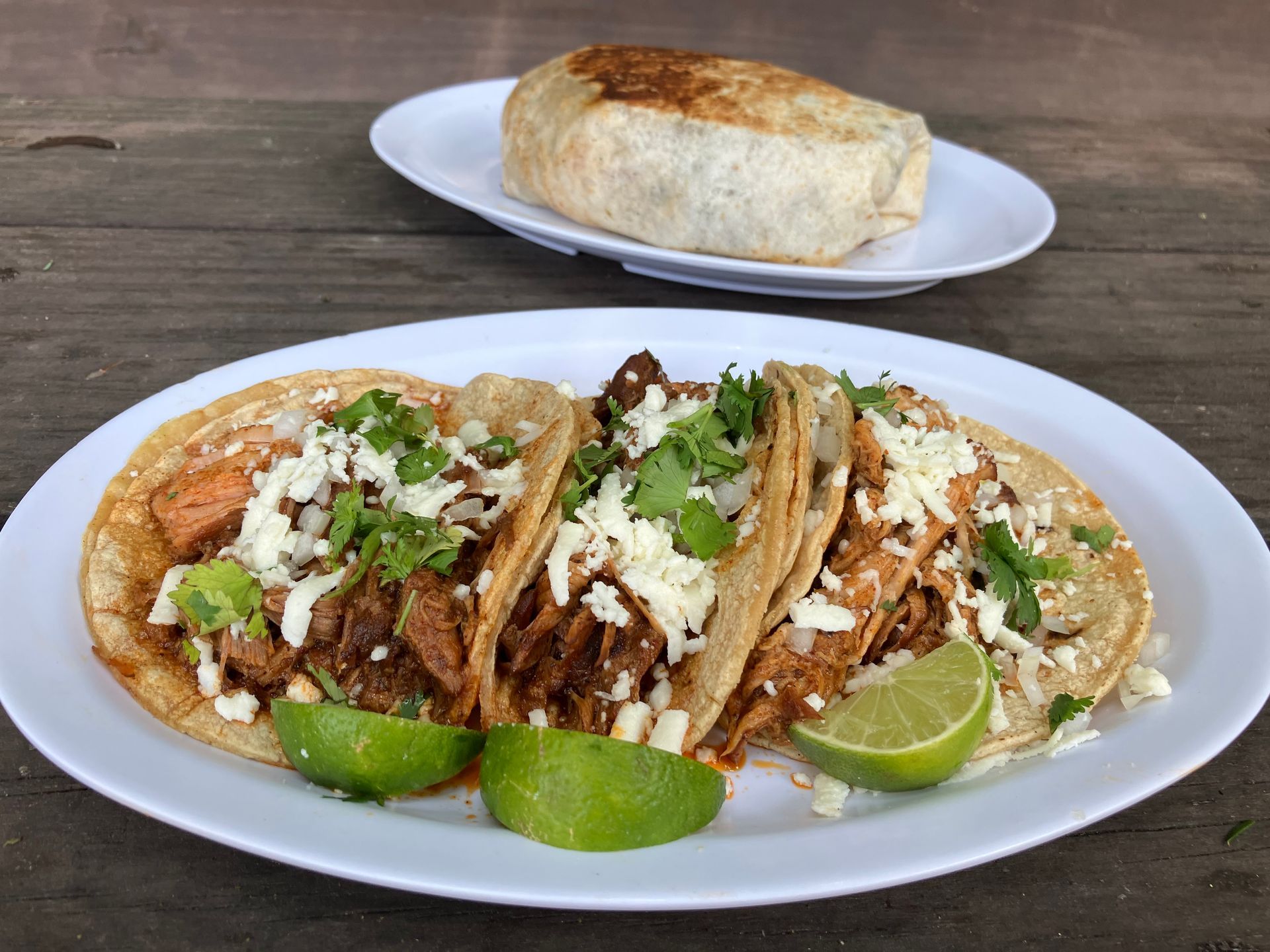 Plate of tacos and a burrito on a white plate on a wooden table, with lime wedges and cilantro.