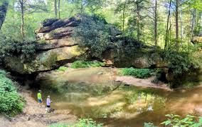 Two people stand near a calm creek, under a large rock formation, surrounded by trees.