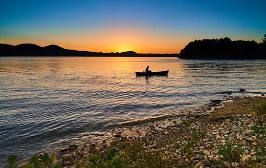 Silhouette of person canoeing on calm water at sunset. Orange and blue sky reflects on the water.