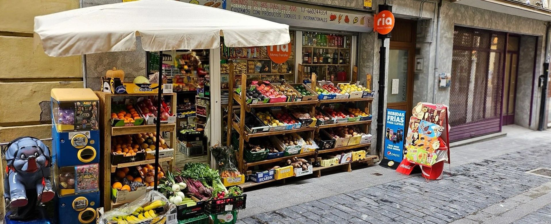 Una pequeña tienda exhibe diversos artículos. Una sombrilla blanca protege algunos artículos a la izquierda, y un farolillo rojo de papel cuelga sobre la entrada.