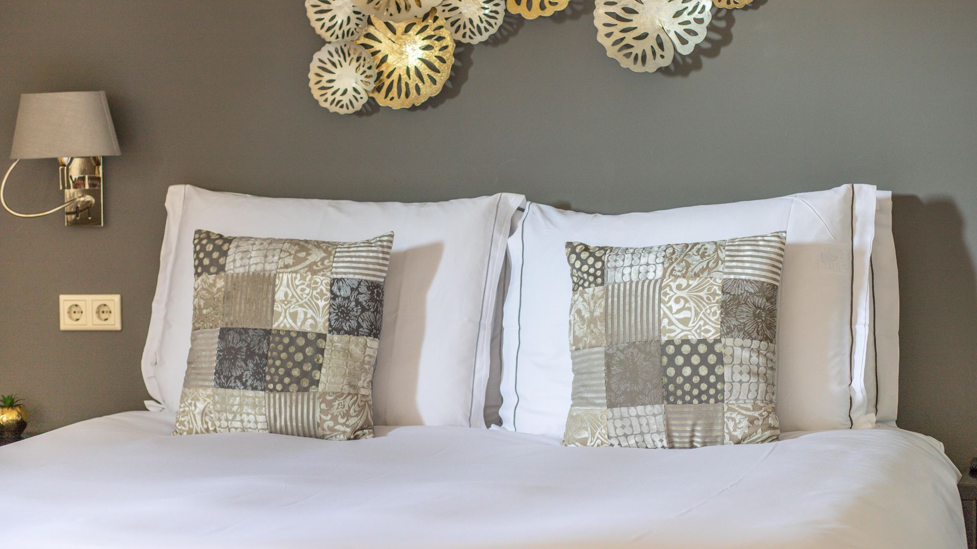 Netjes opgemaakt bed met wit beddengoed, kussens met patroon, een wandlamp en decoratieve muurdecoratie boven het hoofdeinde.