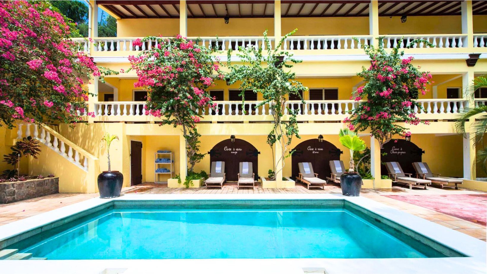 Hotel met een zonnige binnenplaats, een turquoise zwembad, gele balkons en roze bloeiende klimplanten.