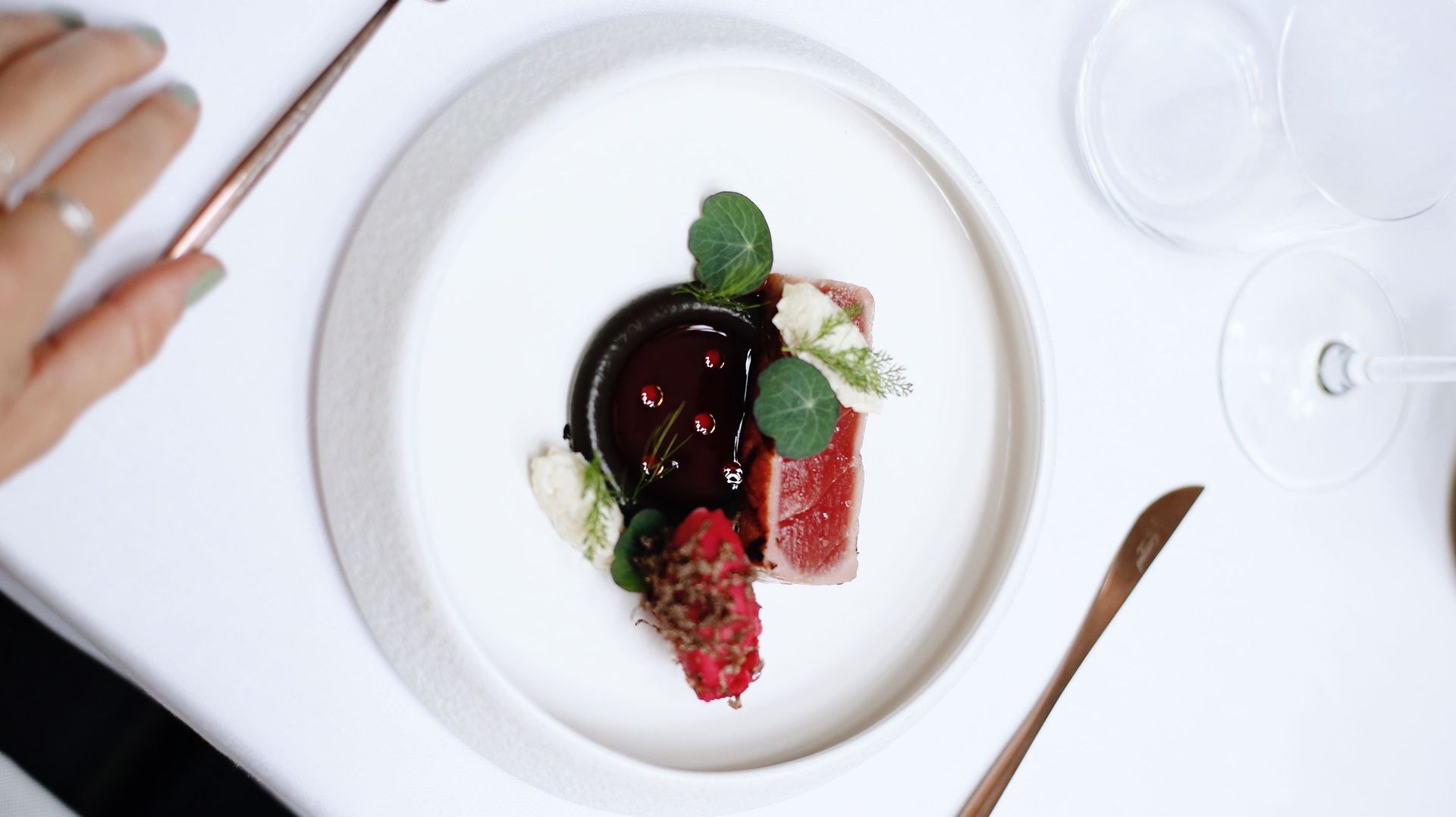 Een dessert op een bord, gegarneerd met chocolade en bessen, staat op een witte tafel, met bestek en een wijnglas erbij.