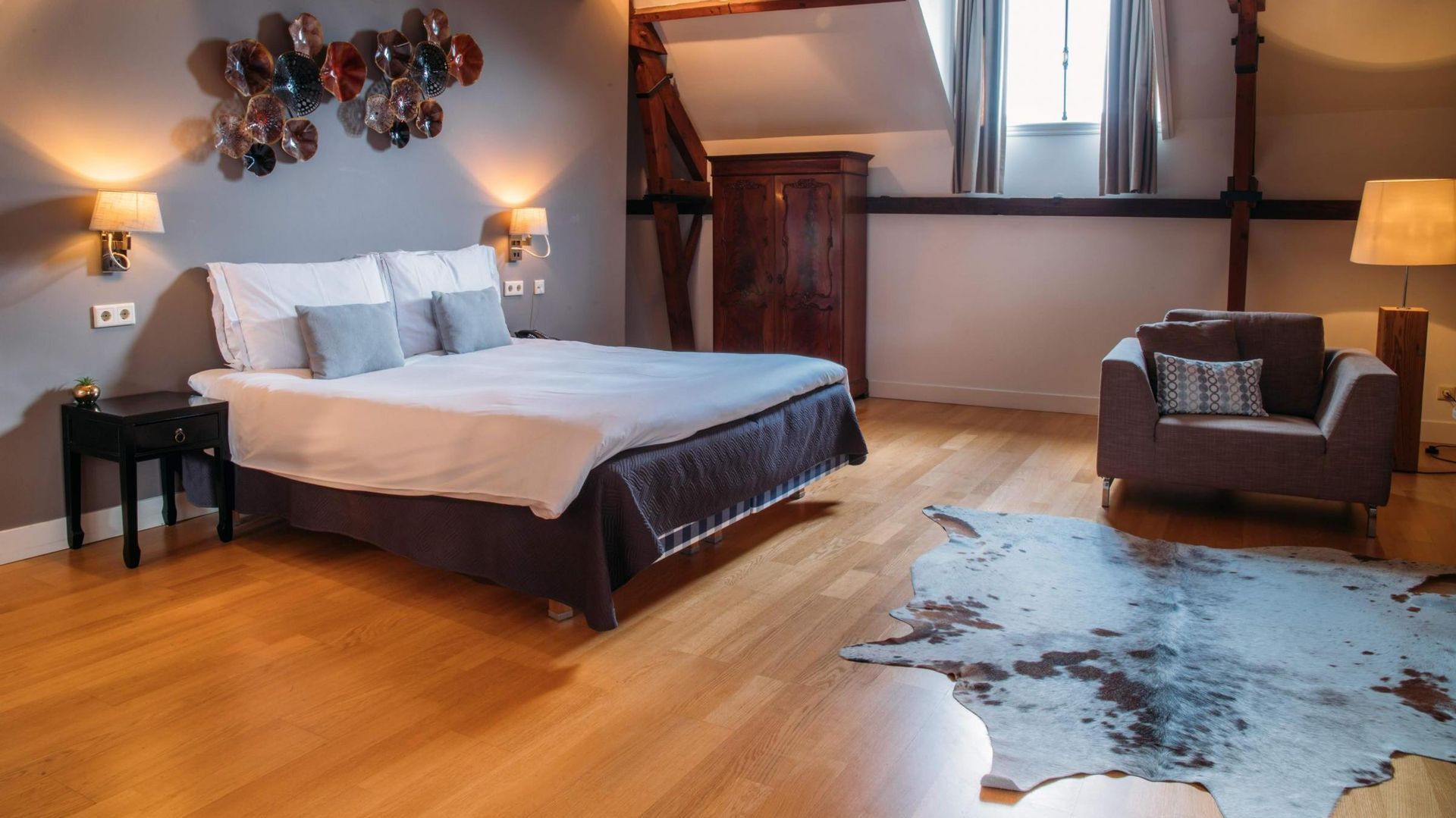 Moderne slaapkamer met wit bed, donkere stoel, houten vloer en koeienhuid vloerkleed.