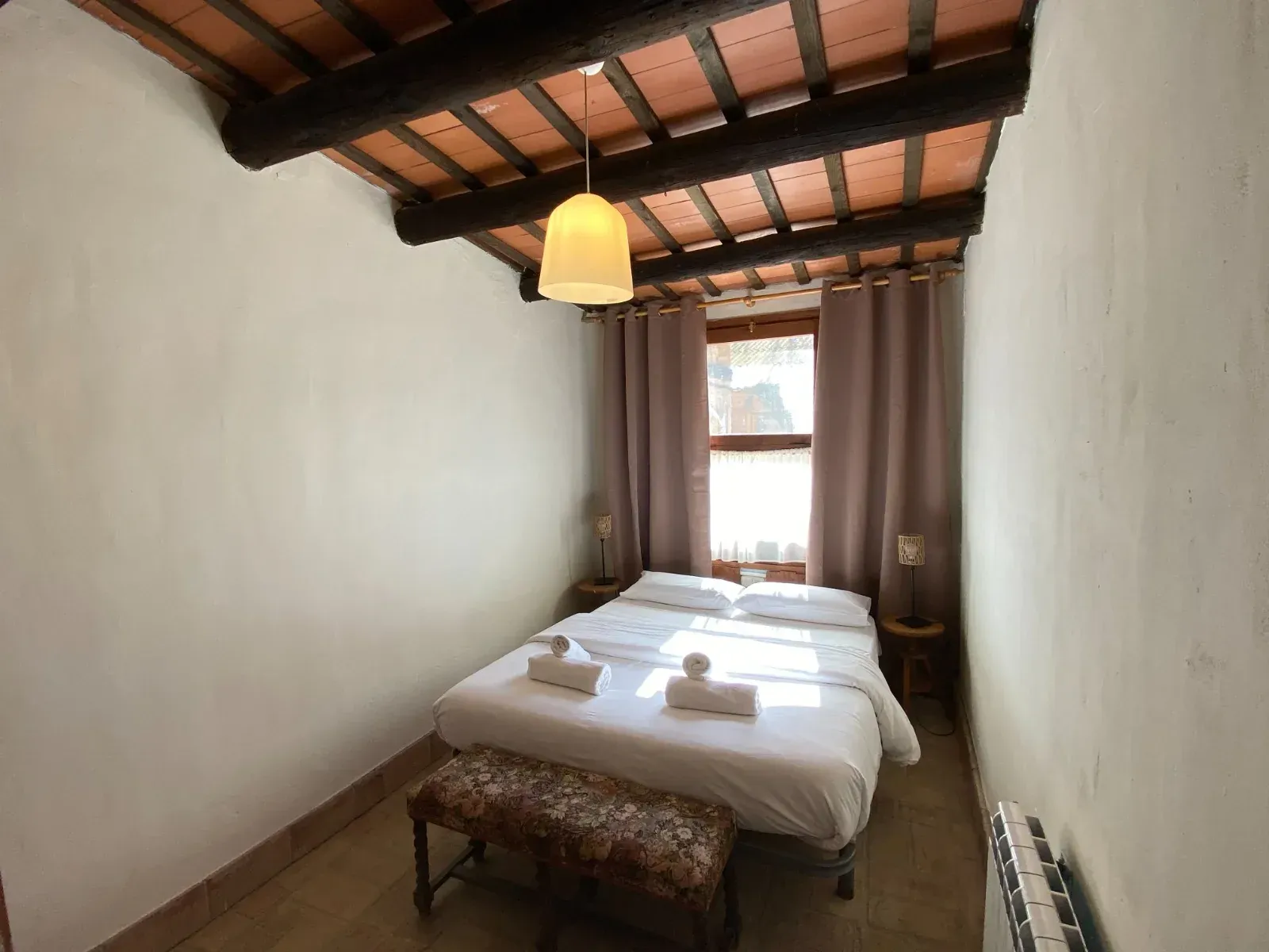 Habitación estrecha con dos camas individuales, paredes blancas, techo de madera y ventana con cortinas.