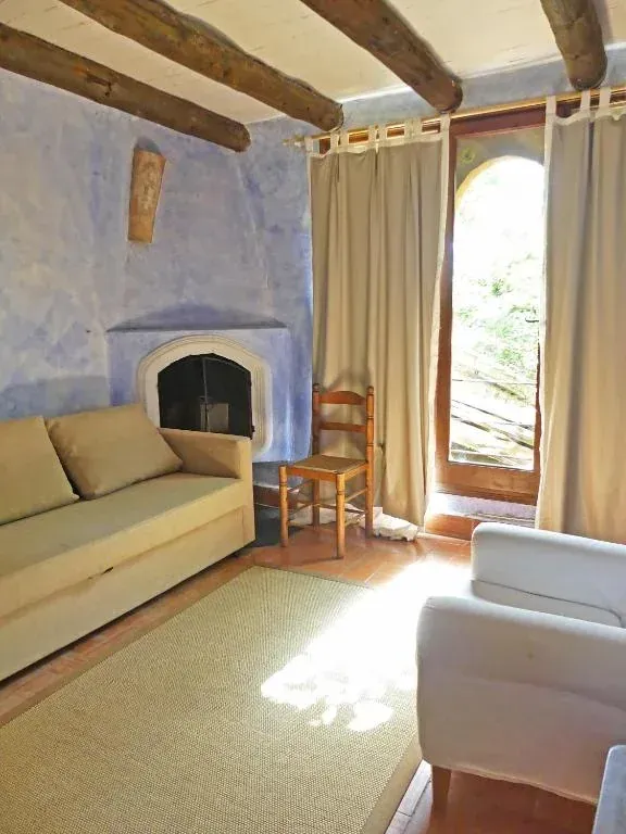 Acogedora sala de estar con sofá beige, chimenea, vigas de madera y luz natural que entra por cortinas altas.