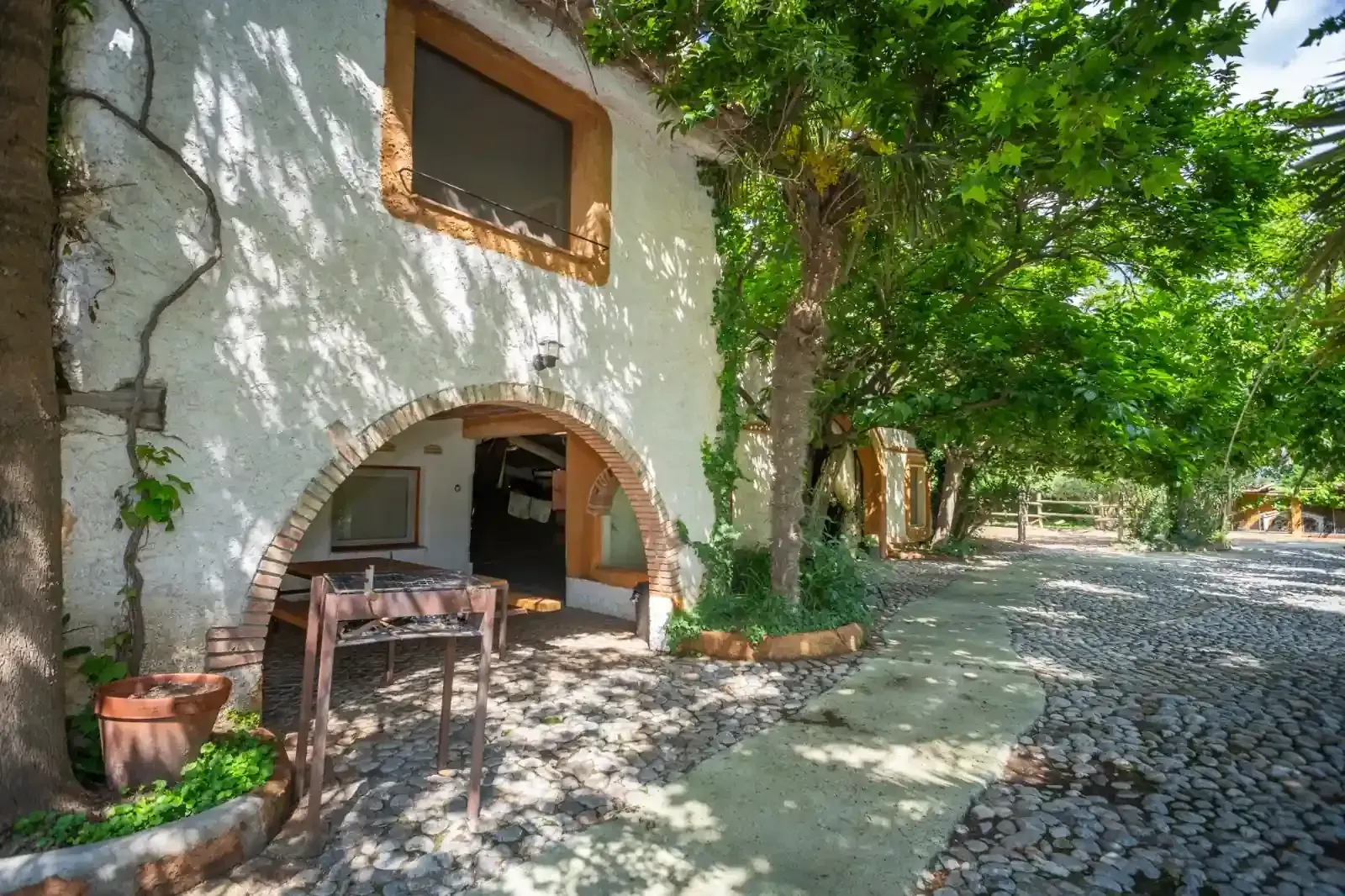 Casa de piedra con patio arqueado junto a un sendero empedrado bordeado de árboles, bañado por la luz del sol tamizada.