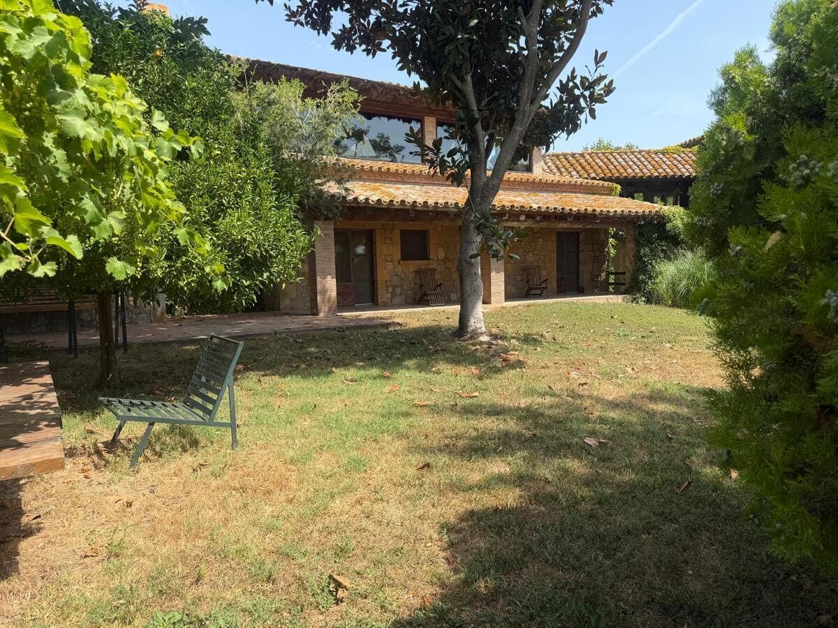 Casa rústica de piedra con tejado de tejas en un patio soleado, enmarcado por árboles y una silla verde.