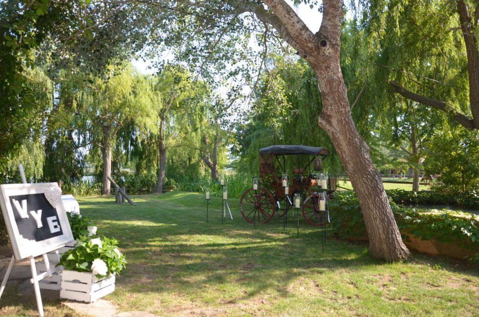 Jardín soleado con árboles, un cartel blanco de boda y una glorieta a la sombra sobre un césped.