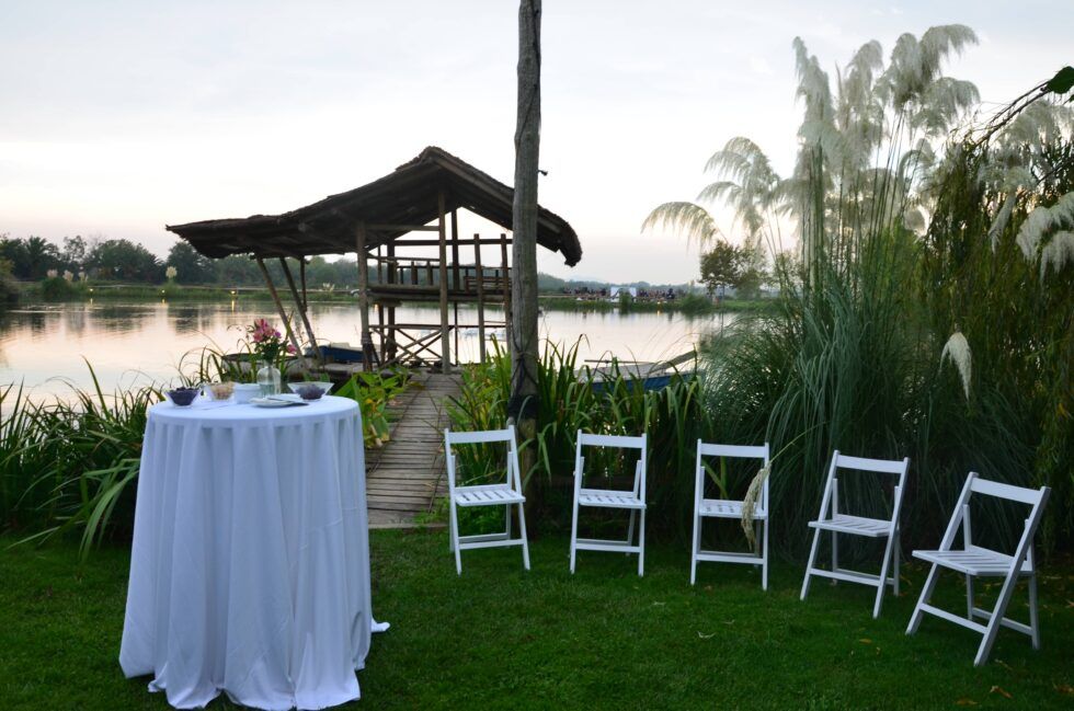 Boda al aire libre junto al lago con sillas blancas y mesa de cóctel al atardecer.