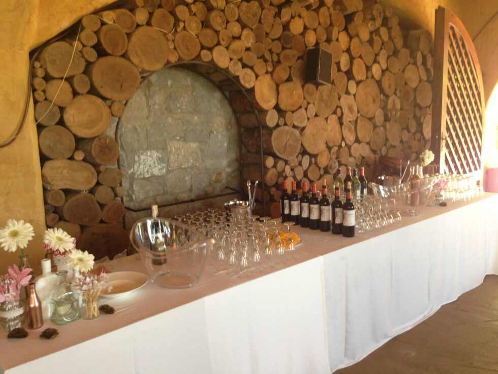 Botellas y copas de vino sobre una mesa junto a una pared rústica de rodajas de madera y un arco de chimenea.