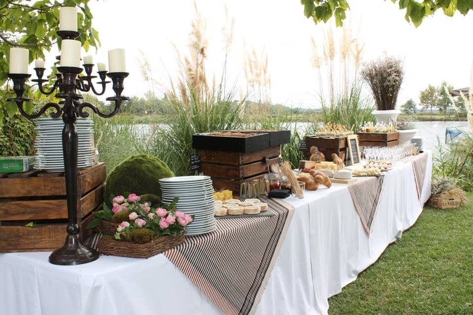 Mesa de buffet al aire libre con manteles blancos, velas, flores, pan y decoración sobre un césped.