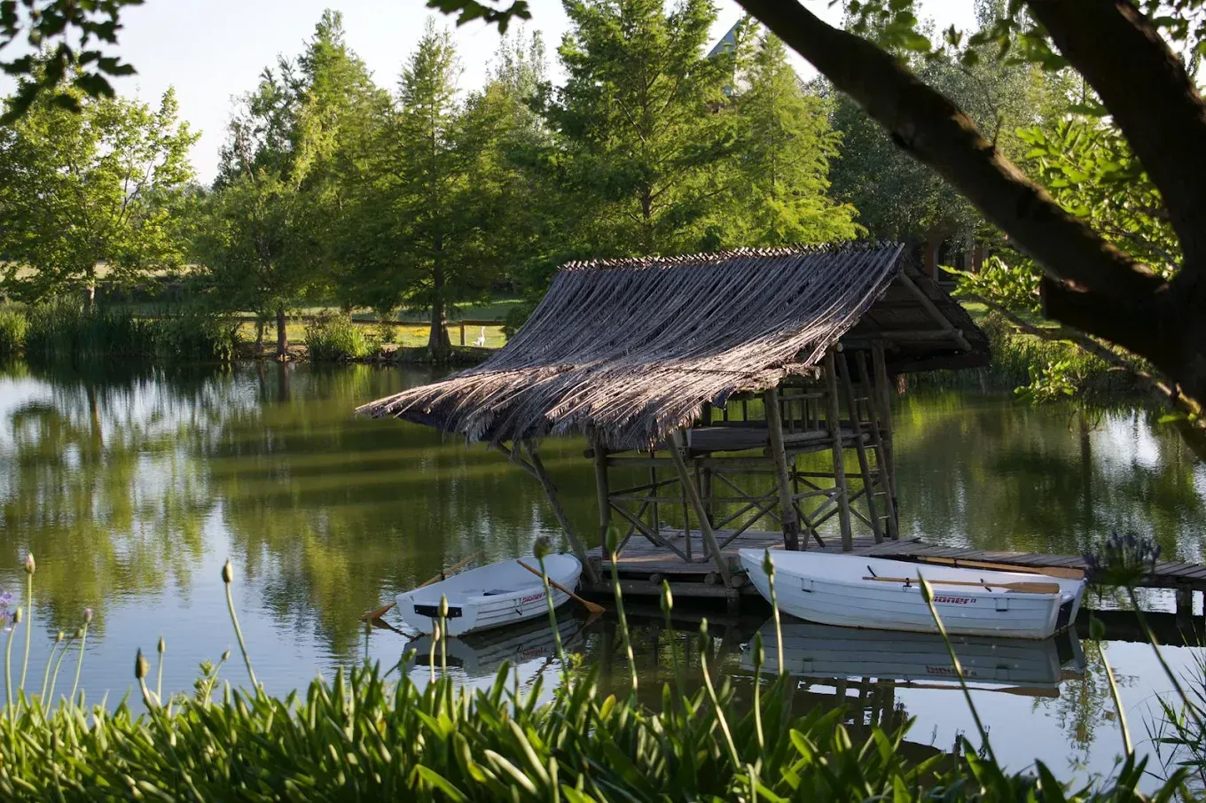 Refugio ribereño con techo de paja y pequeñas barcas amarradas en aguas tranquilas, rodeado de frondosos árboles verdes.