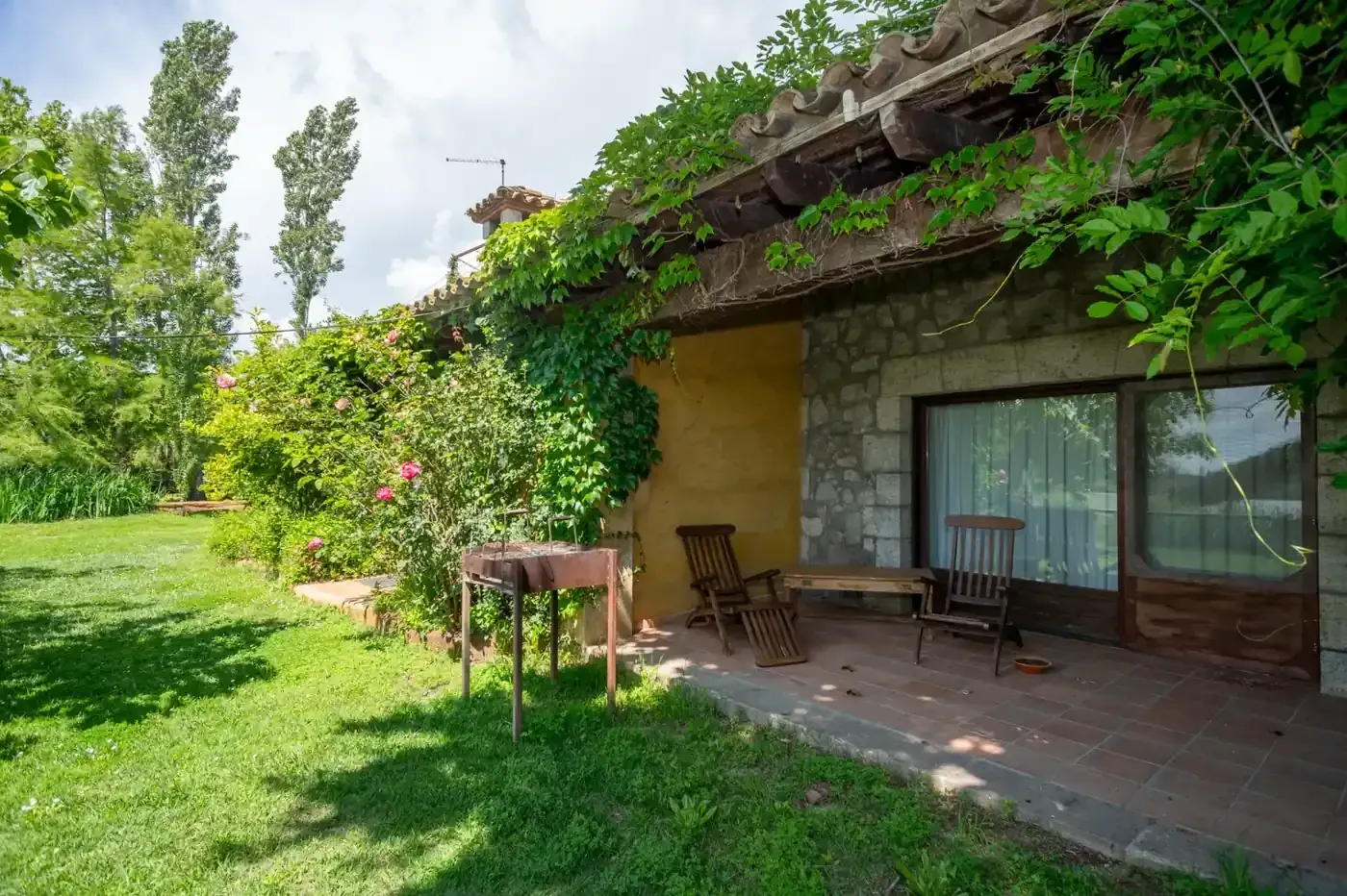 Un patio de piedra con una silla, una mesa y exuberantes enredaderas verdes sobre el tejado, junto a un césped soleado.