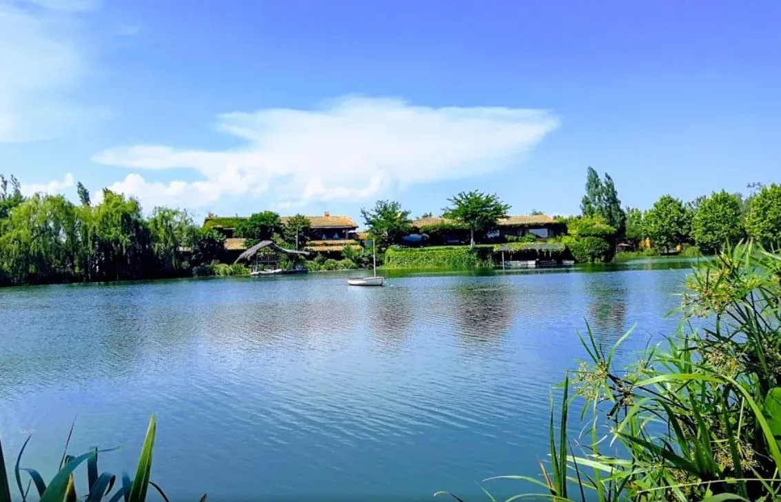 Un lago sereno con aguas ondulantes, árboles verdes y edificios con techos de paja bajo un cielo azul brillante.