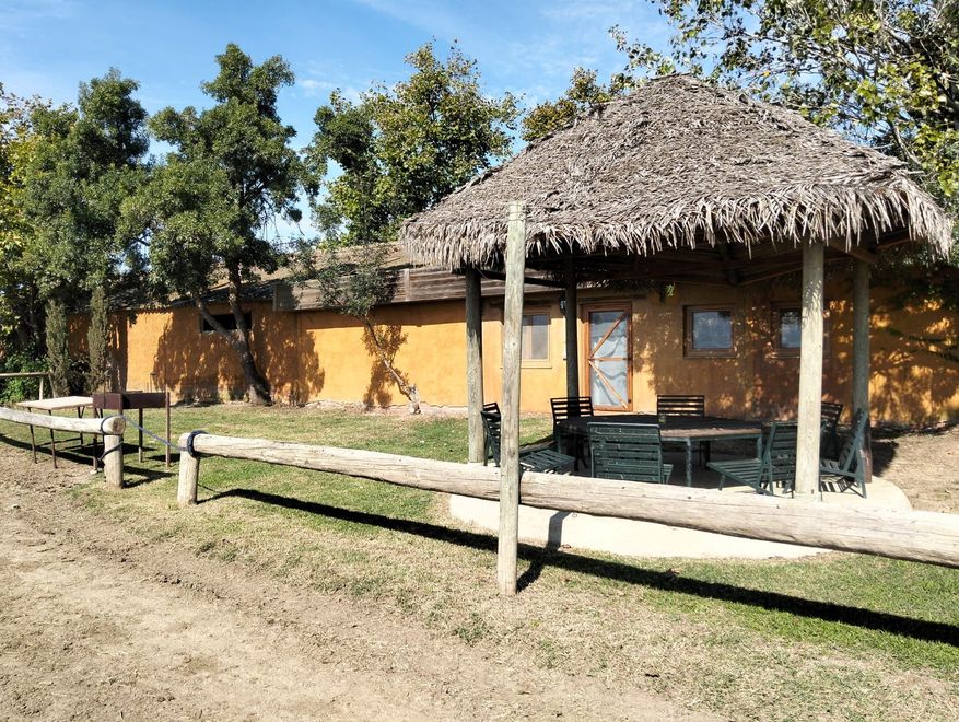 Cabaña con techo de paja junto a una valla de madera en un entorno rural soleado.