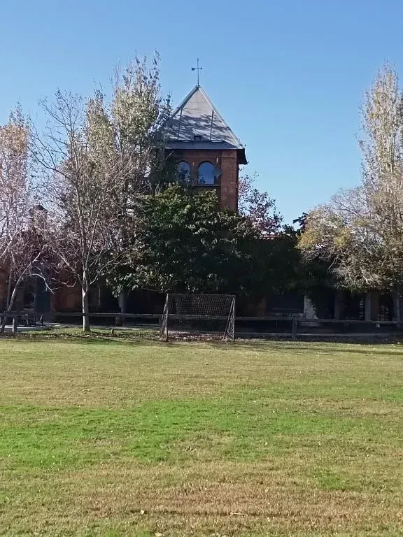 Edificio de ladrillo con tejado a dos aguas detrás de un campo de hierba y árboles bajo un cielo azul claro.