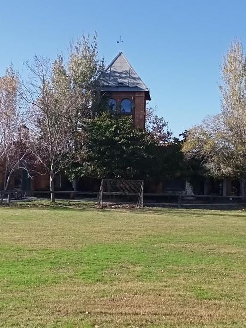 Edificio de ladrillo con una torre central detrás de un campo de hierba y árboles bajo un cielo azul claro.