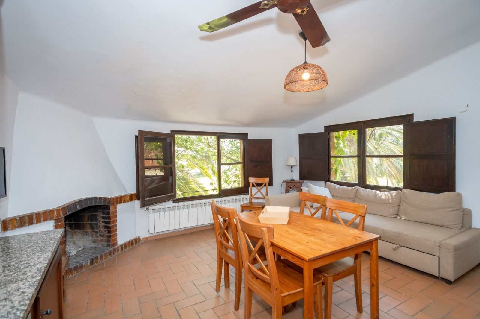 Luminoso salón con mesa de madera, sofá beige, suelo de baldosas, chimenea y ventanas abiertas con contraventanas.
