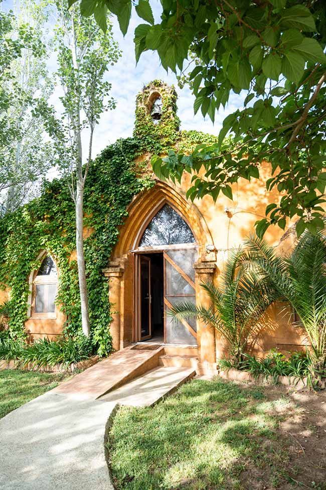 Pequeña capilla cubierta de hiedra con puerta arqueada y sendero de piedra en un jardín frondoso.