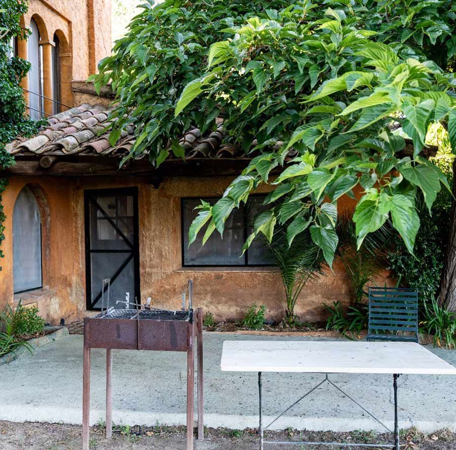 Patio con un árbol frondoso que da sombra a una casita, una mesa de metal y una jardinera.
