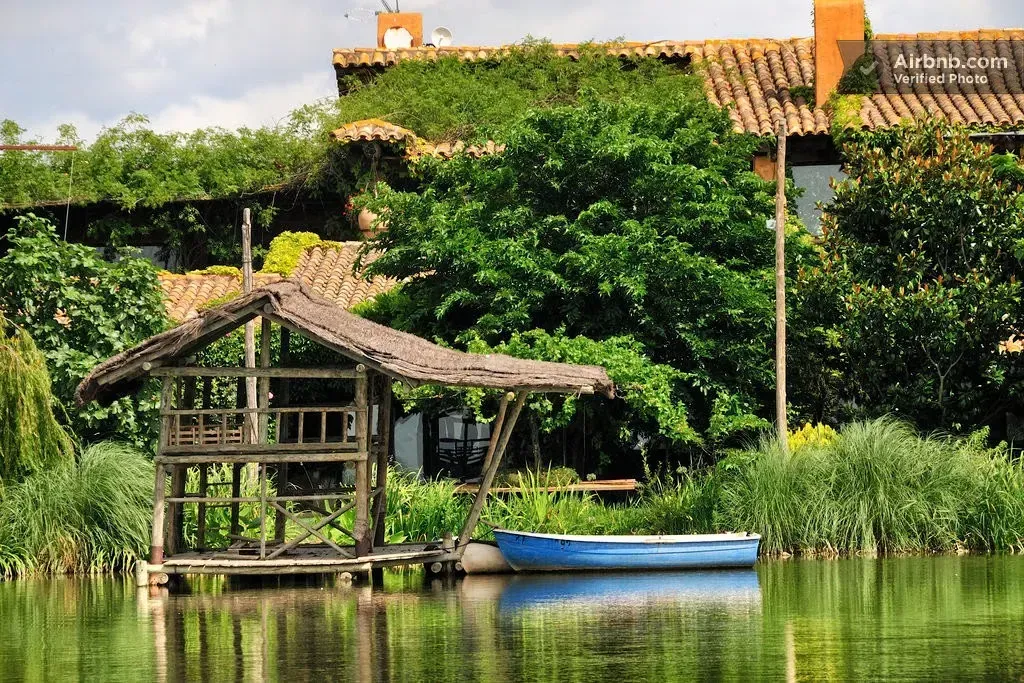 Cabaña con techo de paja a orillas del lago, junto a aguas tranquilas, con frondosos árboles verdes y un barco azul.
