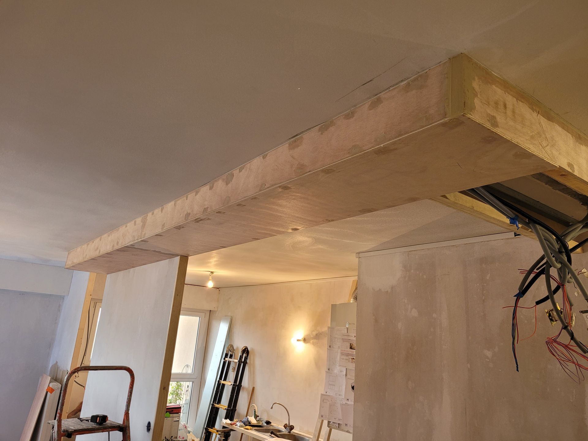 Chantier de construction avec cloison sèche brute sur une poutre de plafond. Câbles et charpente apparents visibles.