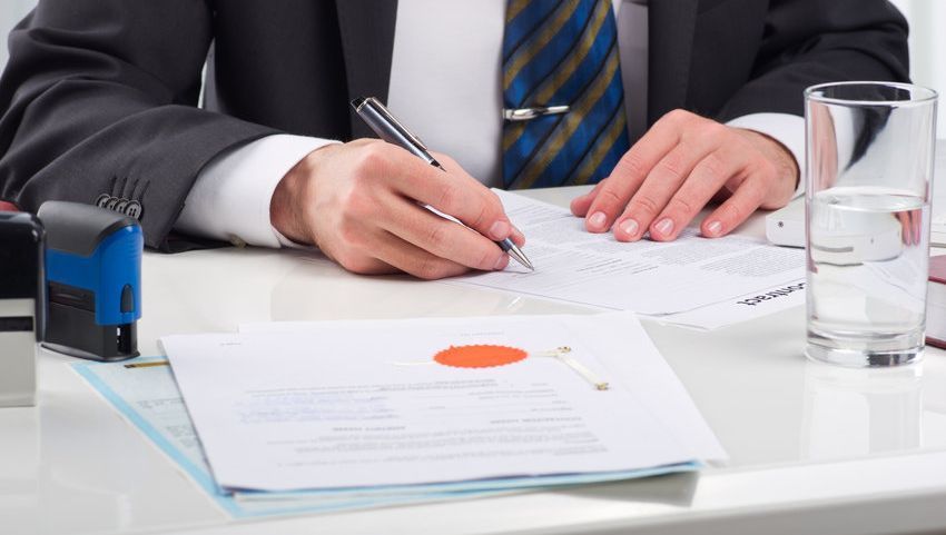 Una persona con traje escribiendo en documentos en un escritorio con un sello, un vaso de agua y un sello oficial.