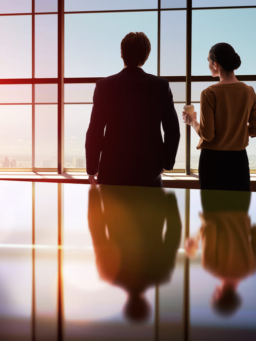 Dos empresarios están de pie junto a un gran ventanal, contemplando la ciudad. Sus reflejos son visibles en la mesa brillante.