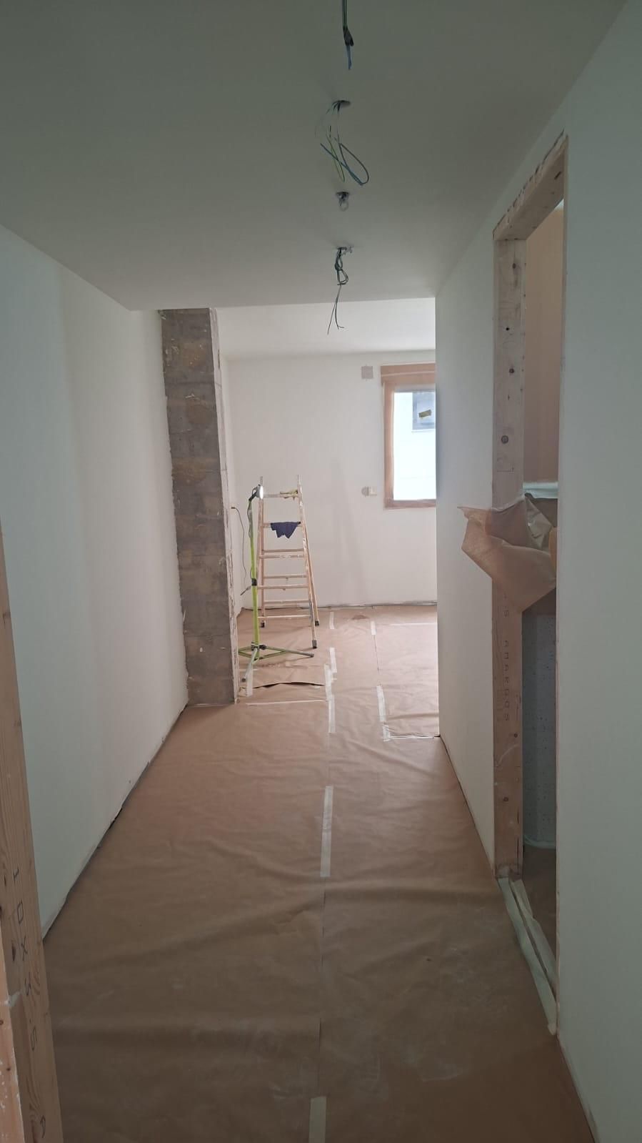 Pasillo renovado con suelo protector, que da a una puerta y una ventana. Paredes blancas, escalera y cables pelados.