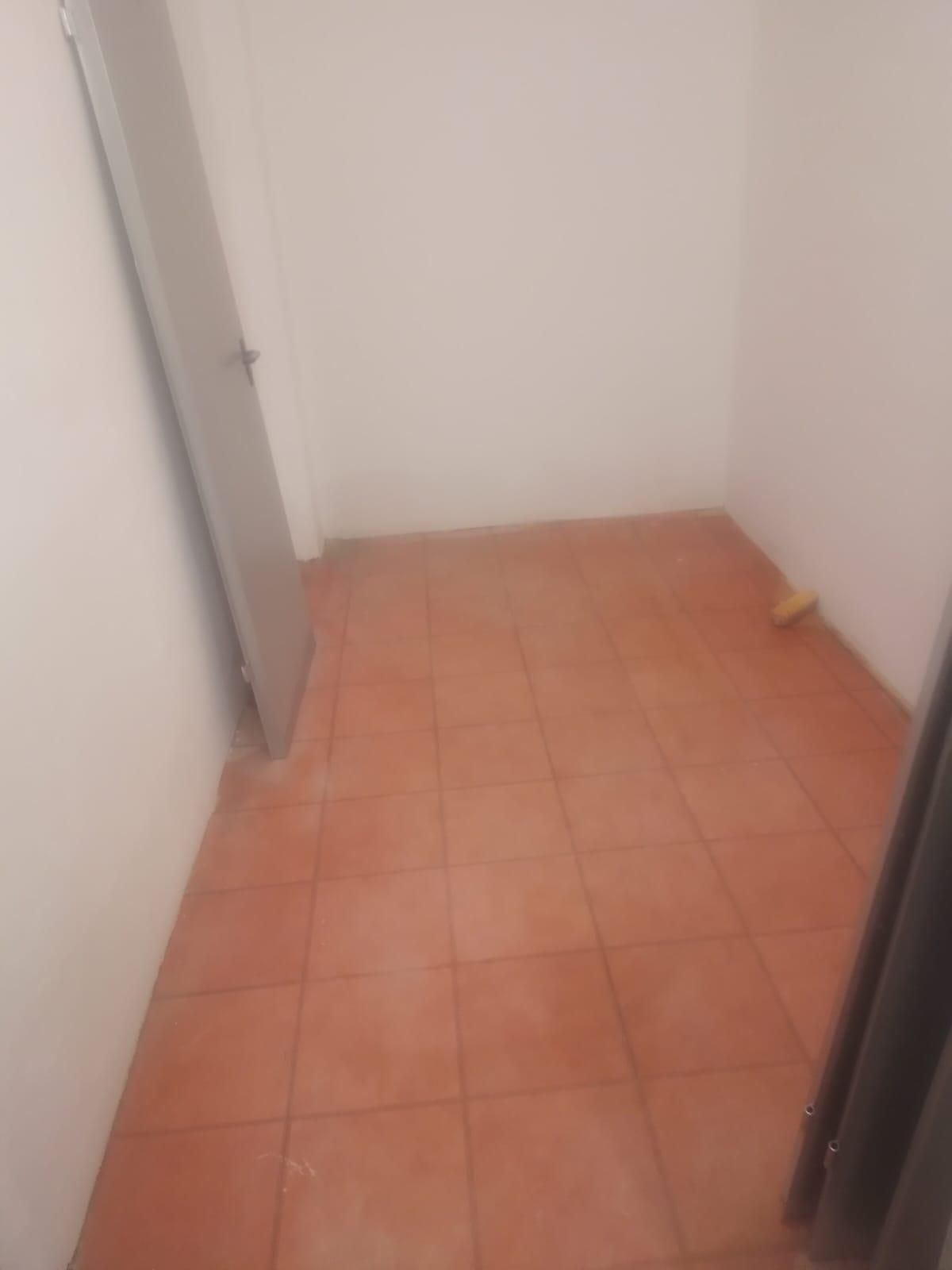 Habitación vacía con suelo de baldosas de terracota, paredes blancas y una puerta gris.