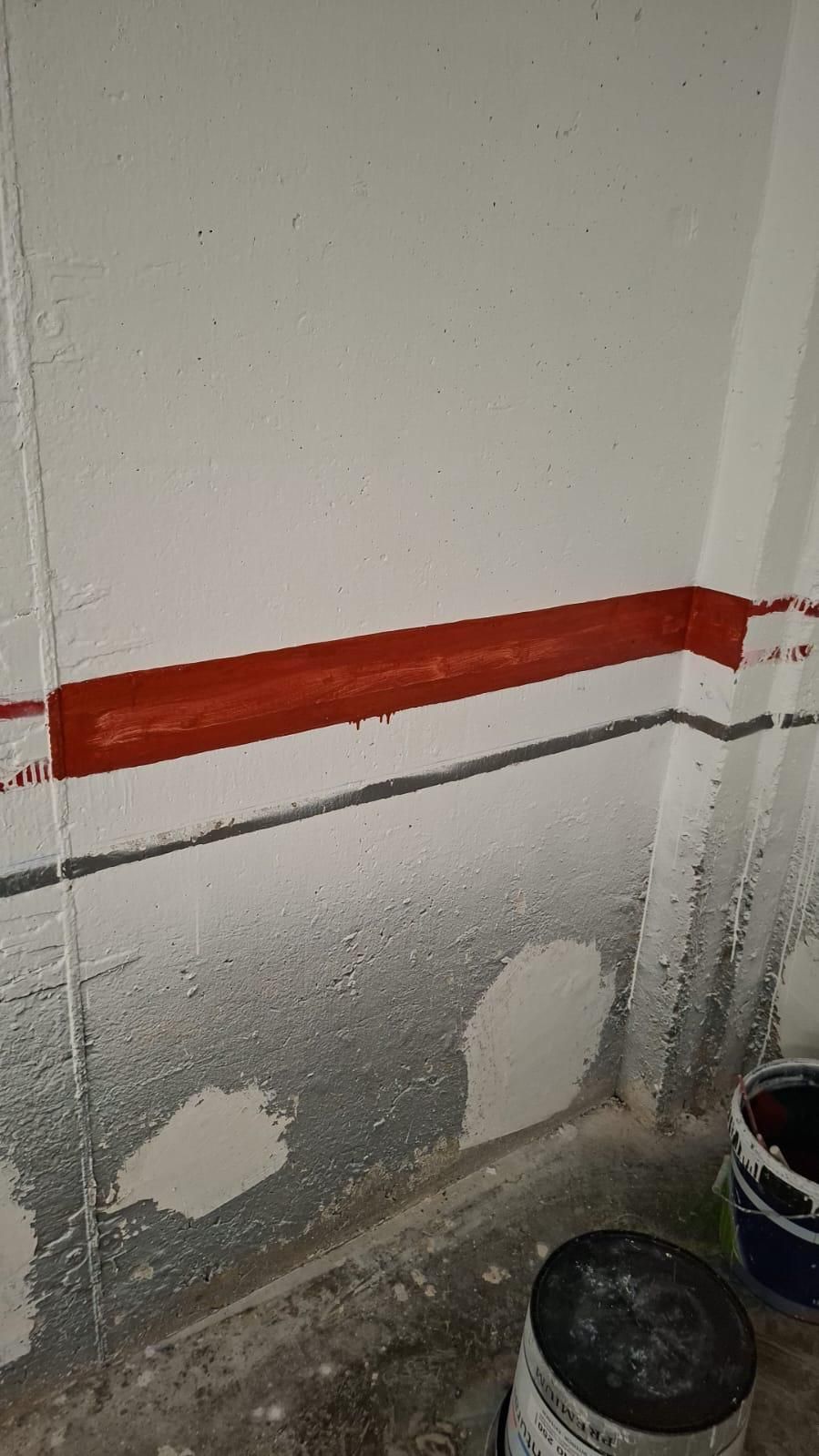 Raya roja pintada sobre una pared blanca, línea gris debajo, latas de pintura en el suelo.