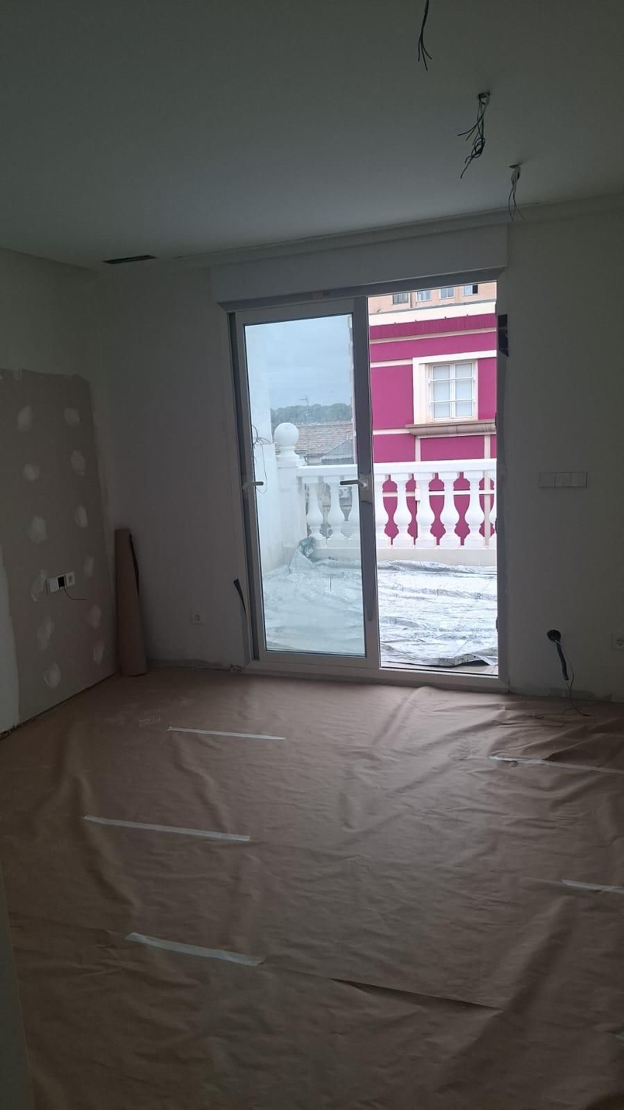 Habitación en construcción, cubierta protectora en piso, puerta corrediza de vidrio, paredes blancas y vista a edificio rosa.