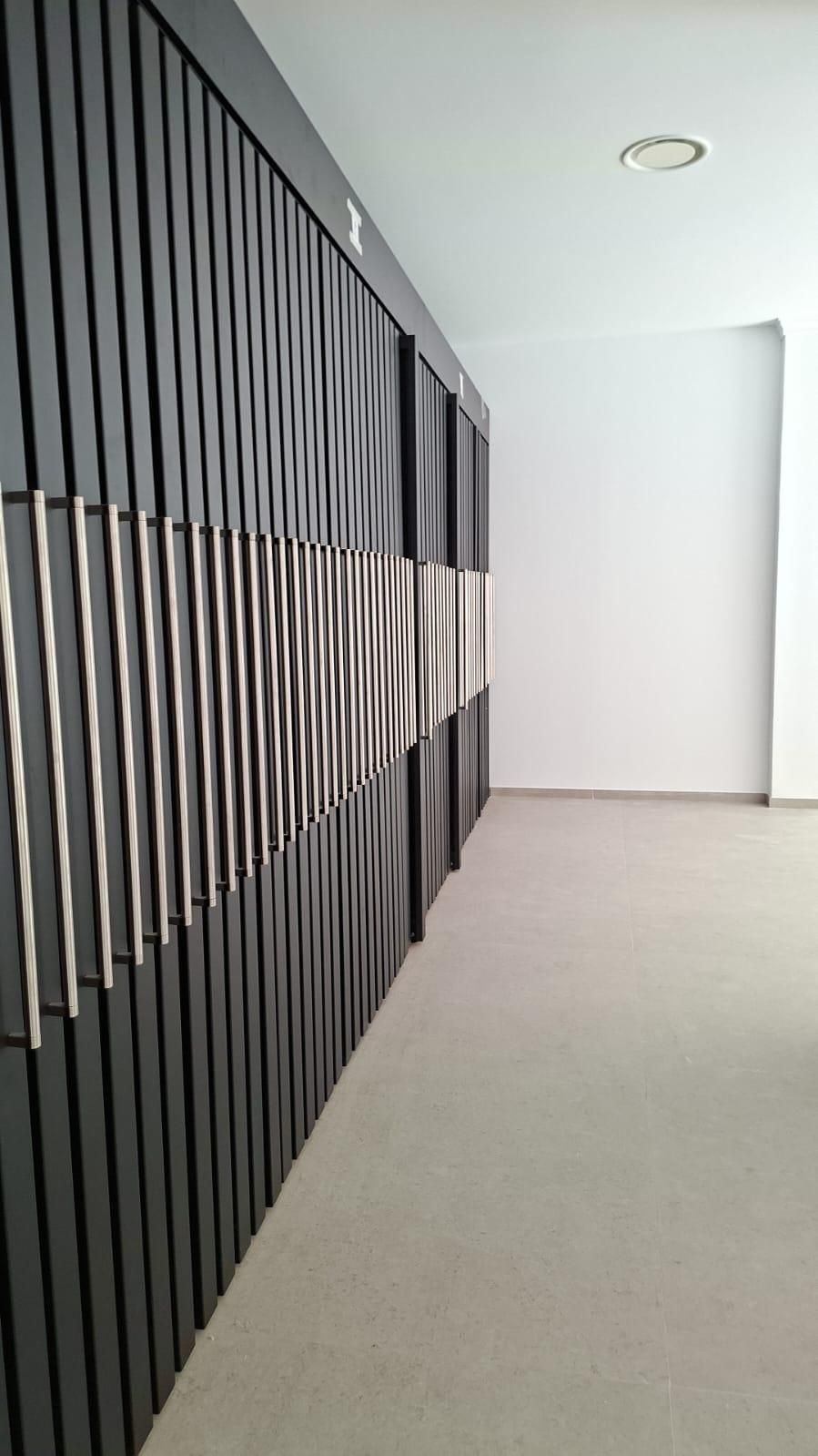 Pared texturizada negra con paneles metálicos ondulados y rectos, al lado de una pared blanca y suelo alfombrado.