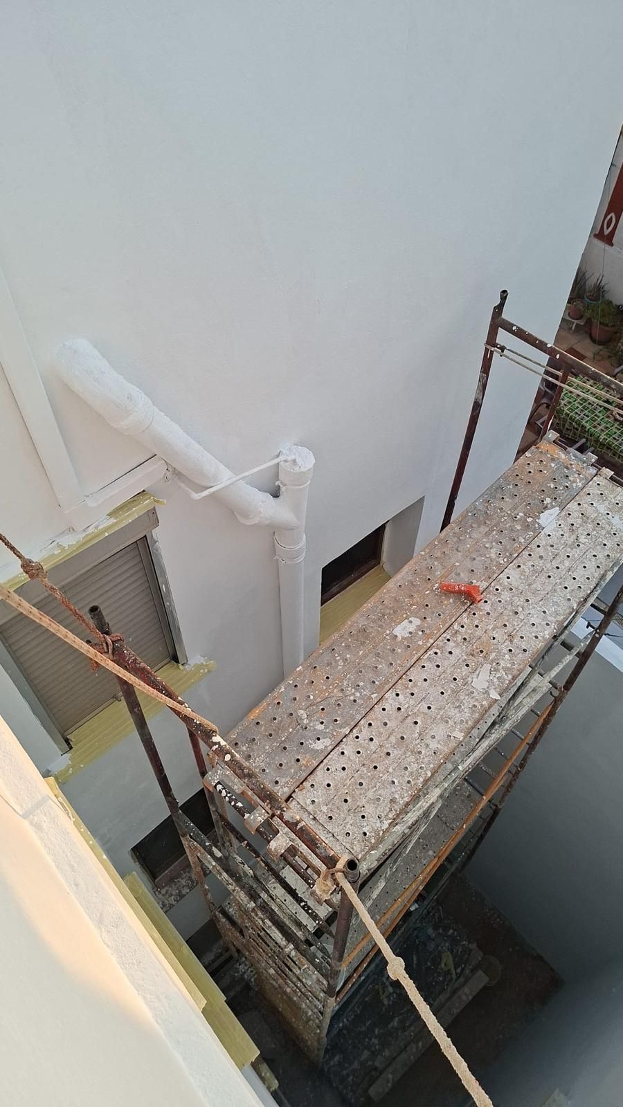 Andamios instalados cerca de la pared blanca de un edificio con canalón. También se ven una ventana y una persiana cerrada.