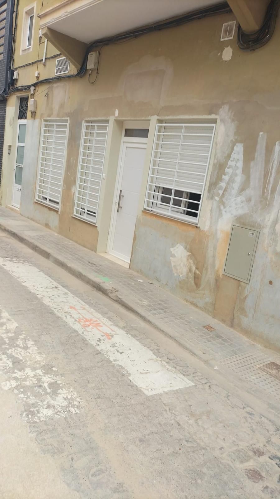 Edificio con ventanas y puerta; acera con pendiente ascendente. Fachada beige, puertas y rejas blancas.