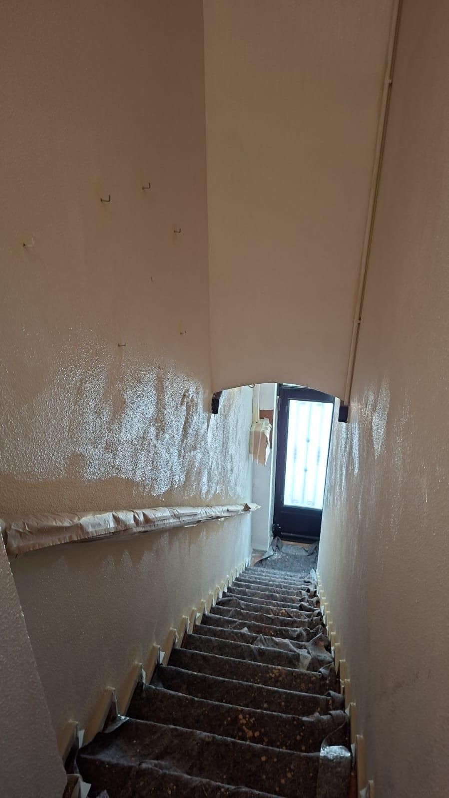 Escalera descendente. Paredes blancas, escalones grises y una puerta al final con una fuente de luz.