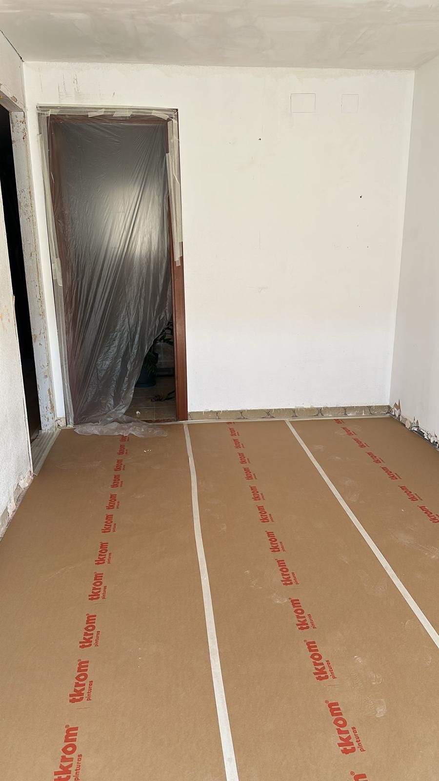 Habitación vacía con suelo de cartón y puerta revestida de plástico. Paredes blancas.
