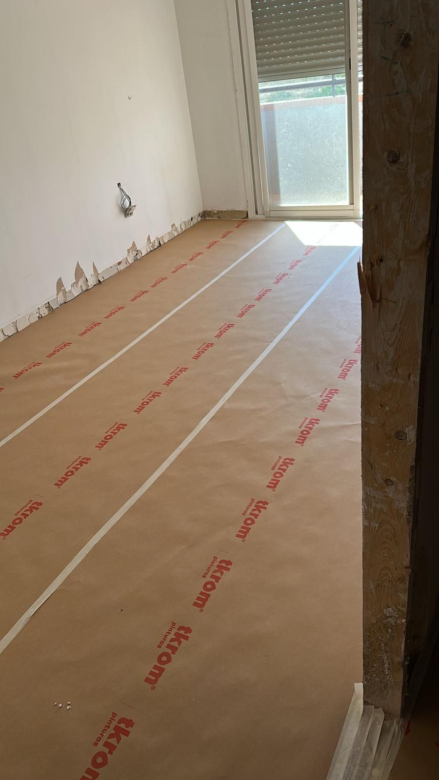 Interior de una habitación con suelo de papel marrón, rayas de cinta blanca y una ventana.