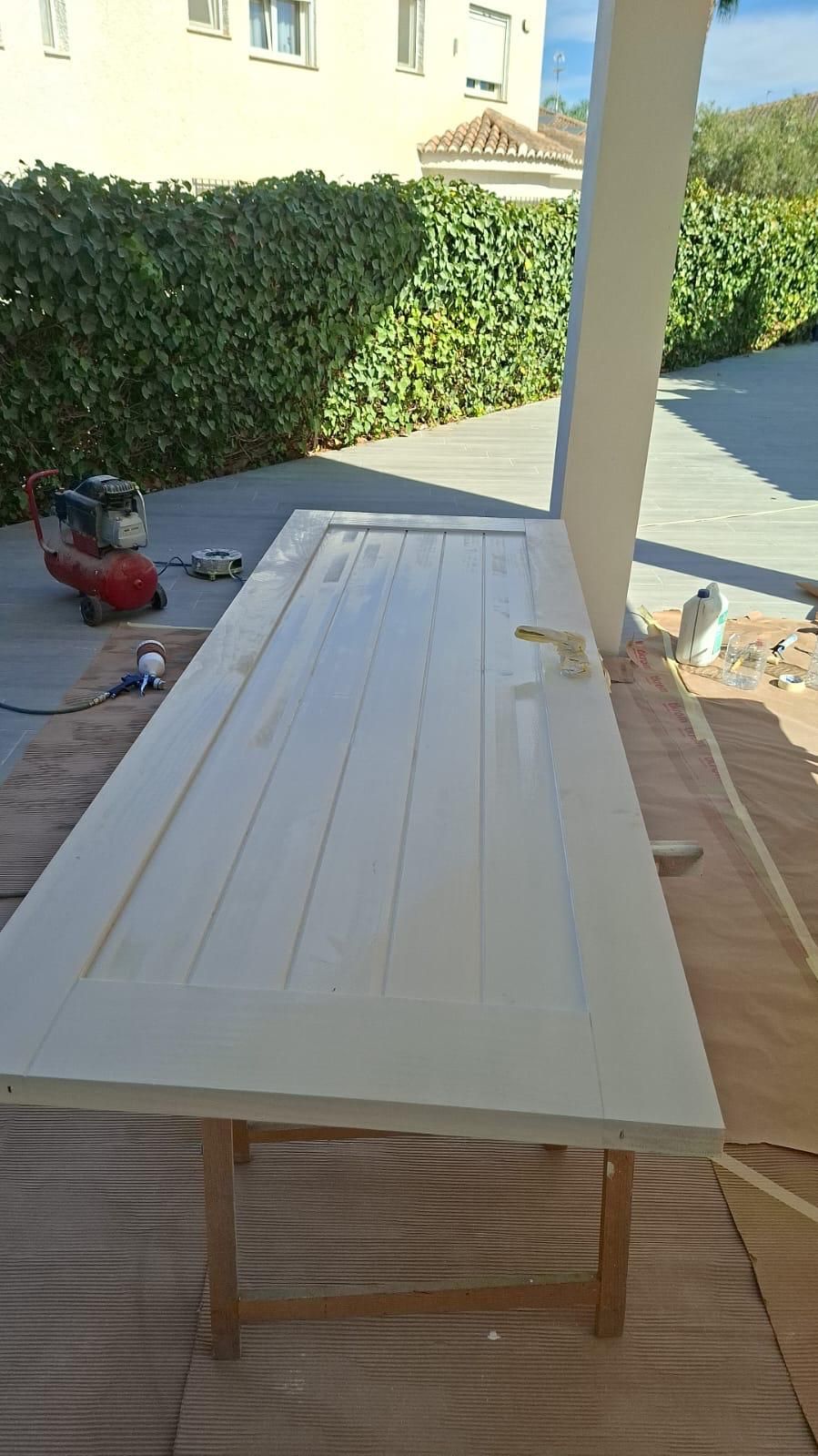 Una puerta pintada de blanco colocada sobre una mesa de madera al aire libre, con un compresor y arbustos en el fondo.