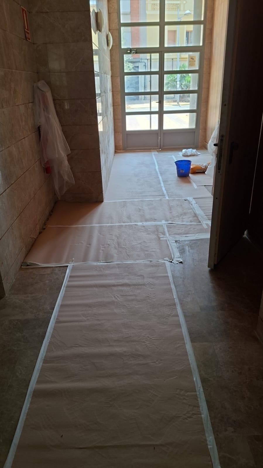 Pasillo en construcción, suelos cubiertos de papel. Puerta al exterior, paredes sin terminar, cubo azul.