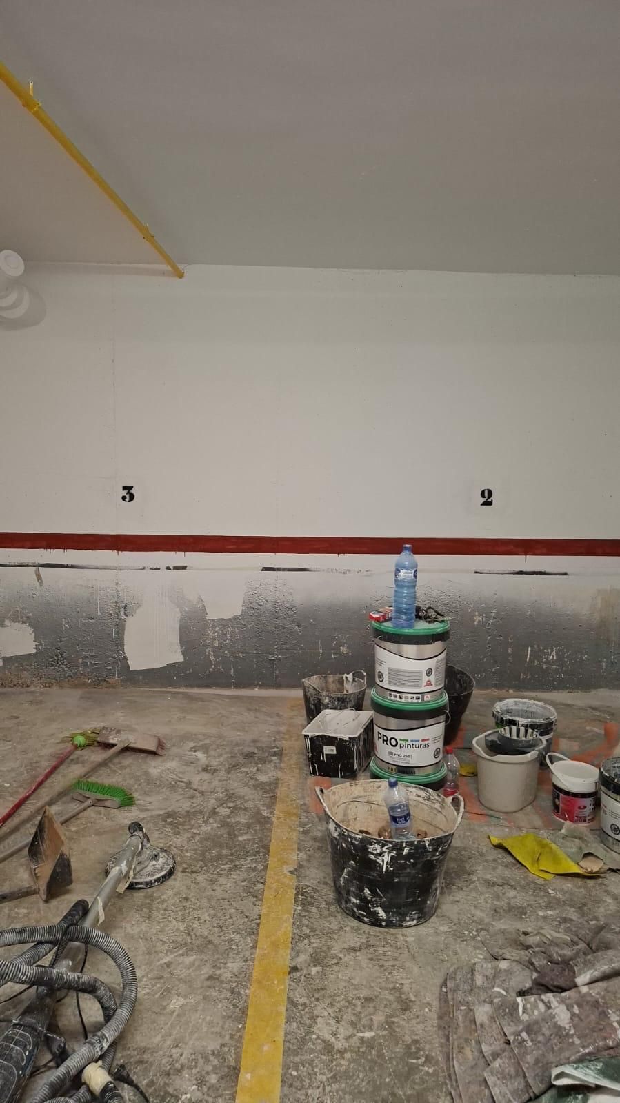 Una habitación que se está pintando; paredes blancas, piso gris, baldes de pintura, botella de agua encima.