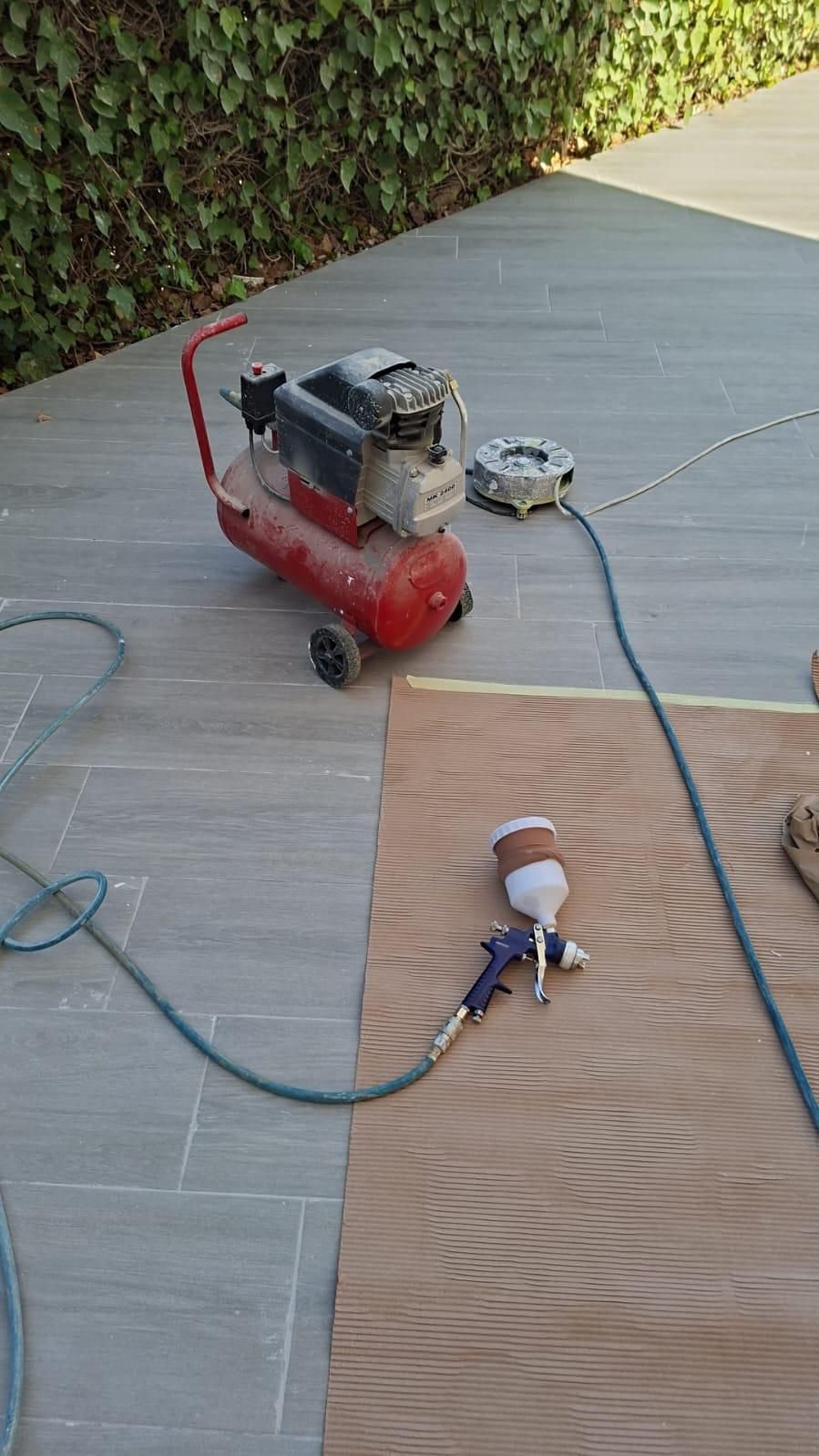Compresor de aire rojo, pistola rociadora y panel de madera sobre una superficie de piedra gris cerca de un seto.