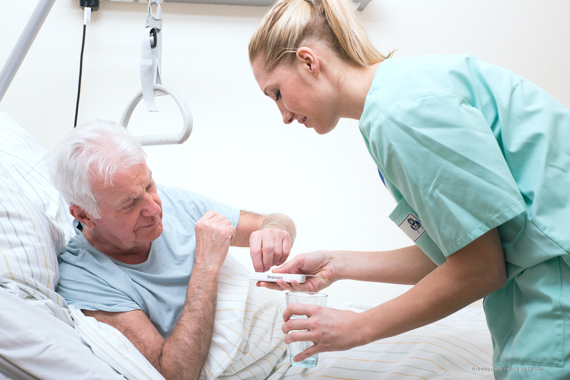 Eine Pflegekraft in grüner OP-Kleidung reicht einem Patienten, der in einem Krankenhausbett ruht, Medikamente
