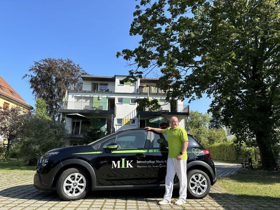 Mann in grünem Hemd neben schwarzem Auto mit "MiK"-Logo vor einem Gebäude.